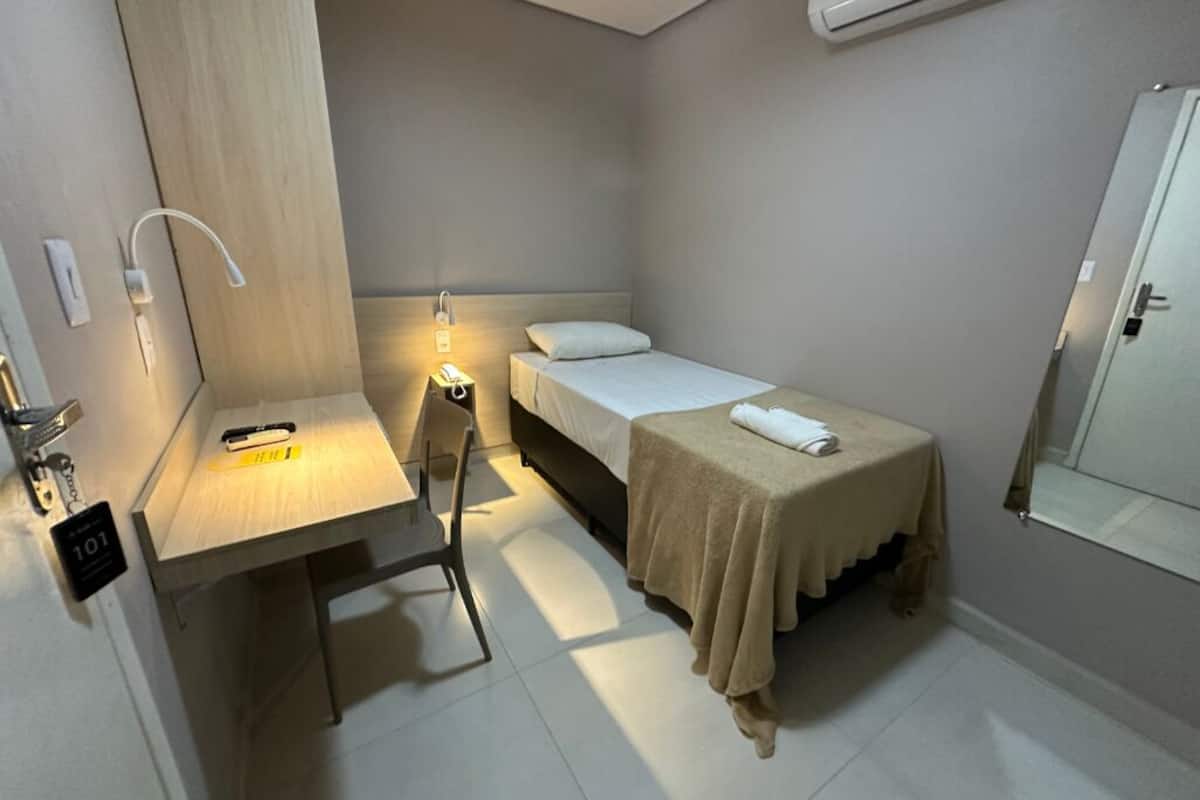 A small, well-lit room with a bed, a desk, and a chair. There is a nightstand with a lamp and a small table with a towel on it.