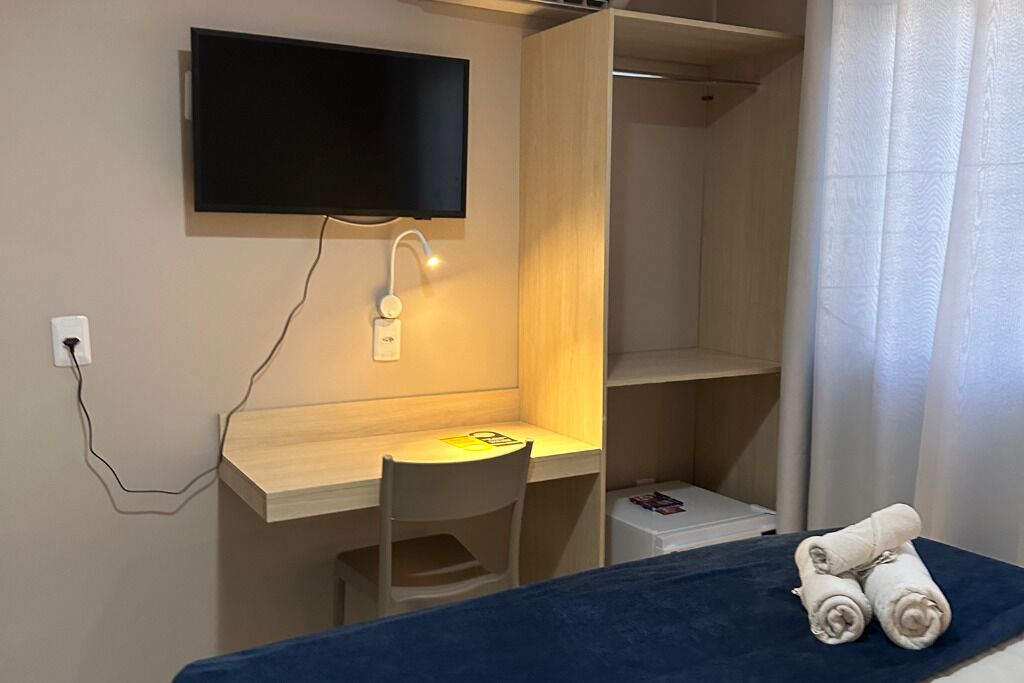A room with a bed, a desk with a chair, a TV mounted on the wall, and a small table with towels.