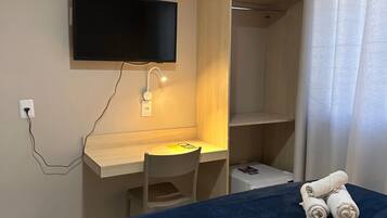 Una habitación con una cama, un escritorio con silla, un televisor montado en la pared y una mesita con toallas.