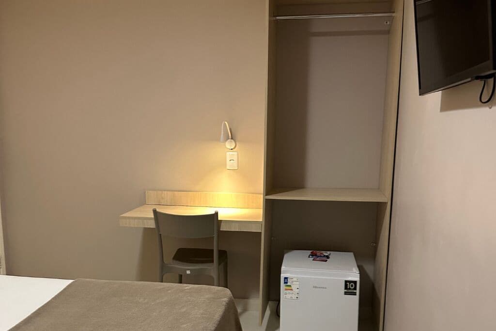 Um quarto pequeno com uma cama, uma escrivaninha, uma cadeira, uma televisão e um frigorífico.