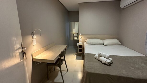 Een kleine hotelkamer met een bed, een bureau en een stoel. Er is een wandairconditioner en een nachtkastje met een lamp.