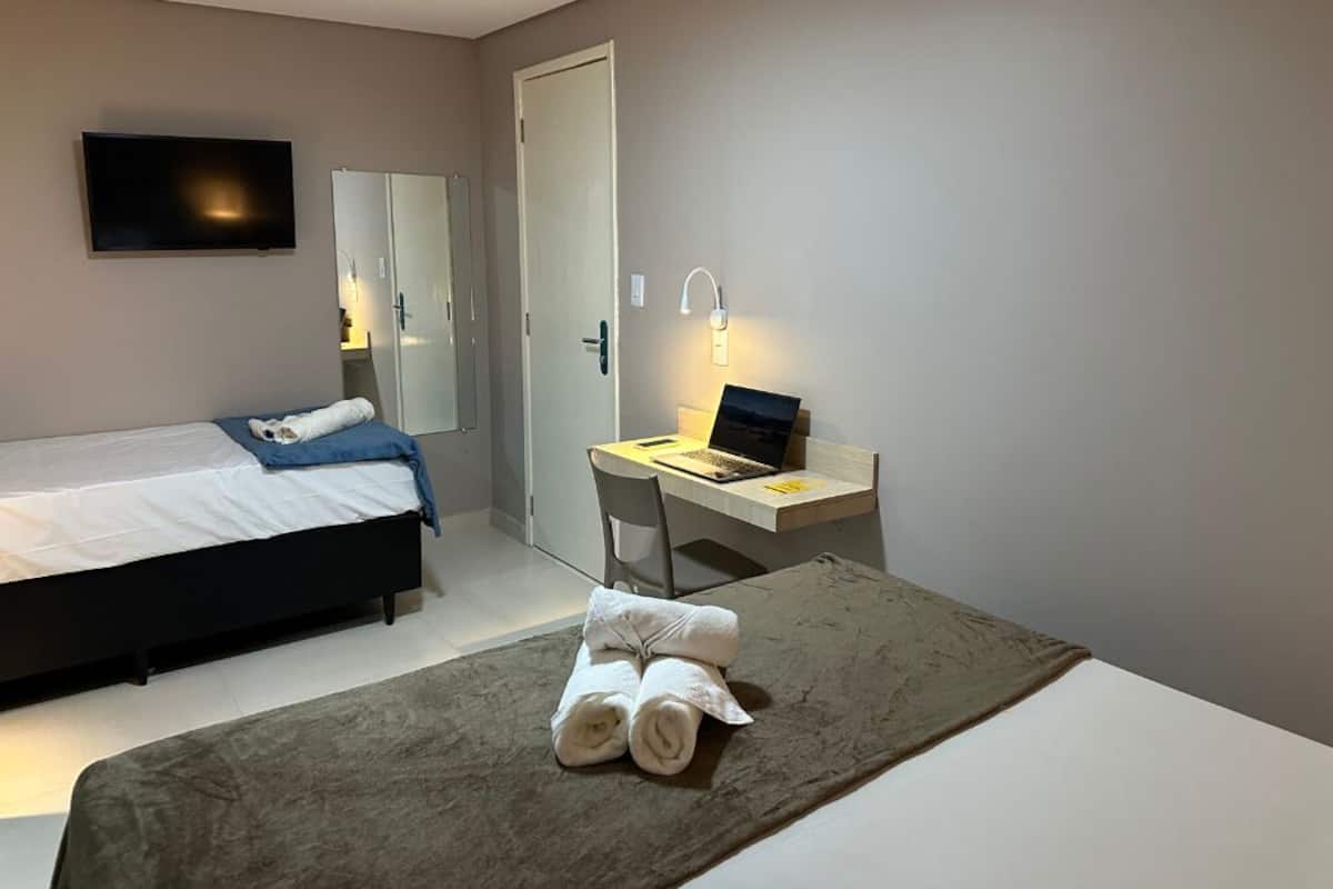 A hotel room with a bed, a desk with a laptop, a TV mounted on the wall, and a bathroom door.
