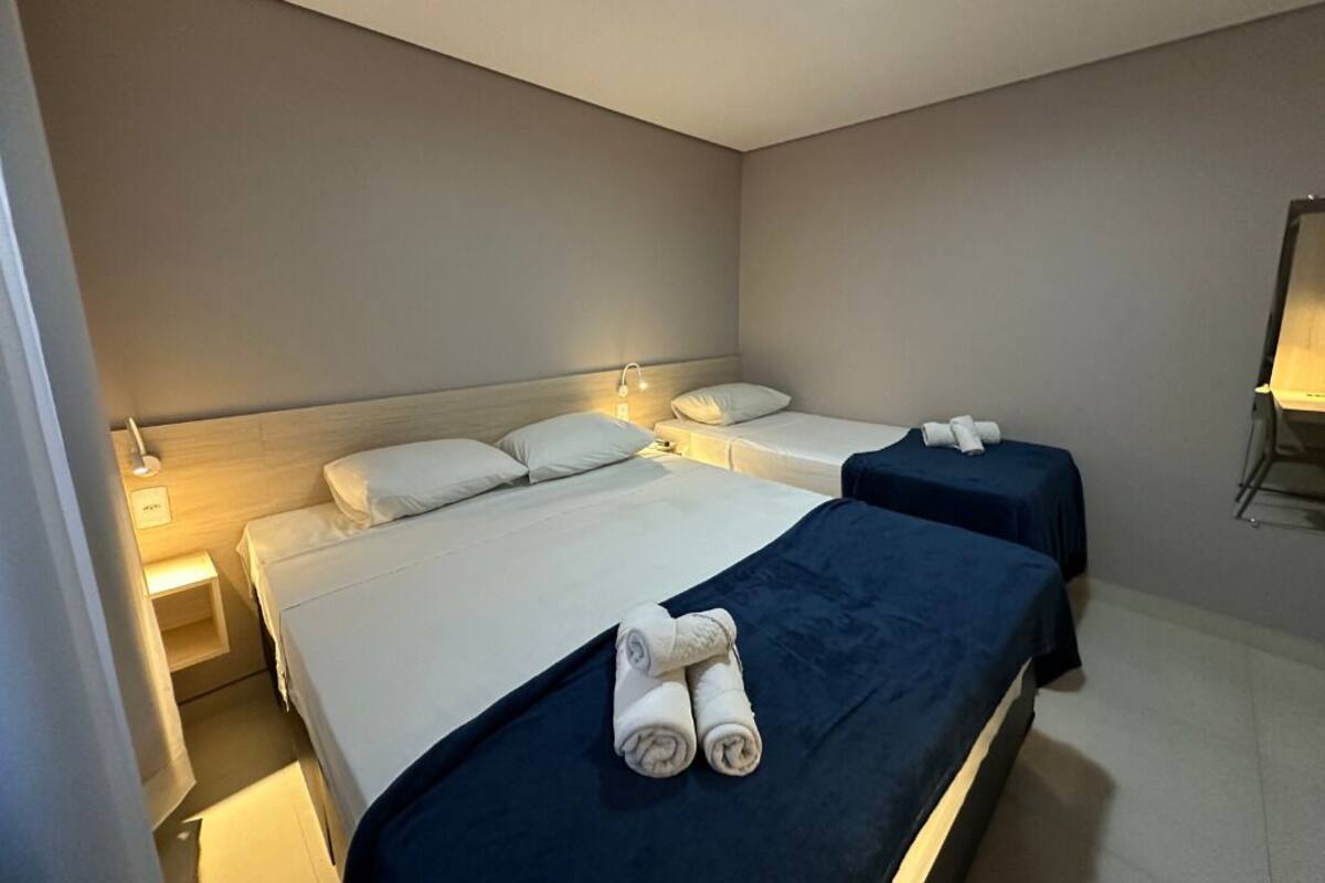 A hotel room with two beds, each with white linens and a dark blue bedspread. There are two bedside tables with lamps, and a small table with a lamp in the corner.