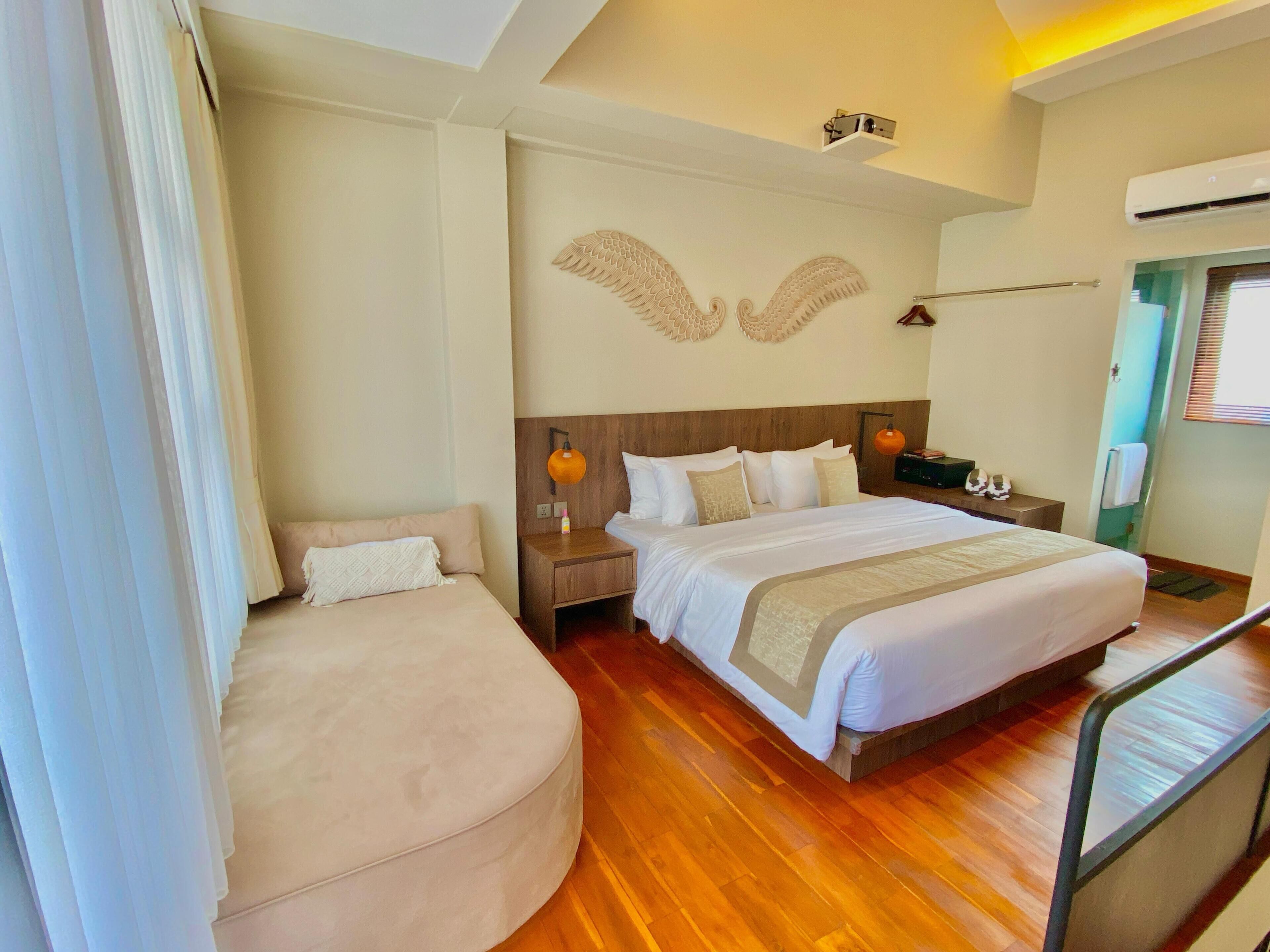 A hotel room with a large bed, a smaller seating area, a wooden floor, a wall-mounted air conditioner, and a ceiling light fixture.