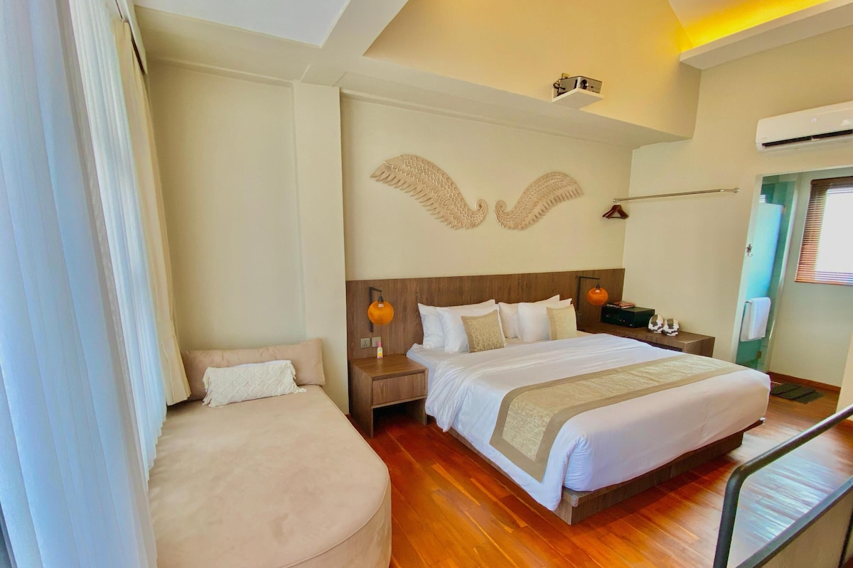Une chambre d’hôtel avec un grand lit, un petit coin salon, un plancher en bois, un climatiseur fixé au mur et un luminaire au plafond.