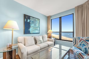 A modern living room with a beige sofa, two lamps, a glass coffee table, and a large window with a view of the beach. - Unit 0702E a beautiful 2 bedroom and 2 bath unit (ORANGE BEACH)