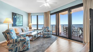 A living room with a sofa, two armchairs, a coffee table, and a ceiling fan. Large windows offer a view of the ocean. - Unit 0702E a beautiful 2 bedroom and 2 bath unit (ORANGE BEACH)