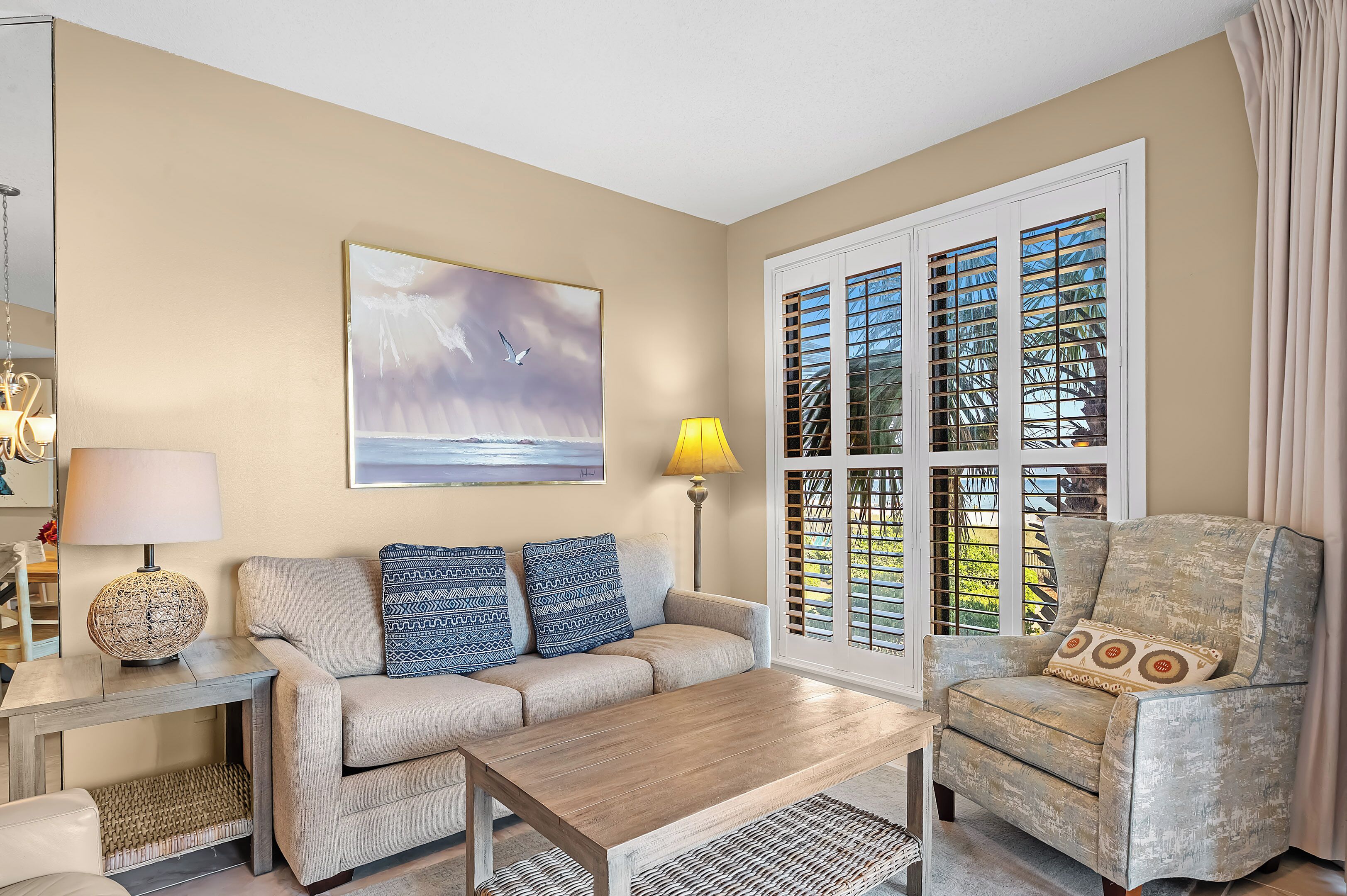 A living room with a beige sofa, a wooden coffee table, a patterned armchair, a painting of a seascape, and a window with shutters.