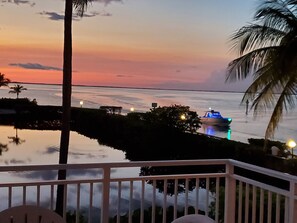 A sunset view from a balcony with a railing, palm trees, and a body of water. - Waterfront Key Largo Escape. Spacious 2-bedroom condo, heated pool, boat slip. (Key Largo)