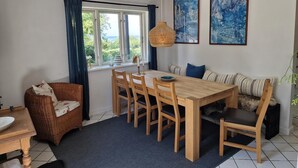 Ein Essbereich mit Holztisch und Stühlen, einem Korbstuhl, einer Couch mit gestreiften Kissen und einem großen Fenster mit Vorhängen.