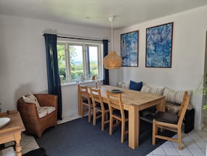 A dining area with a wooden table and chairs, a wicker chair, a sofa with striped cushions, and a large window with curtains.