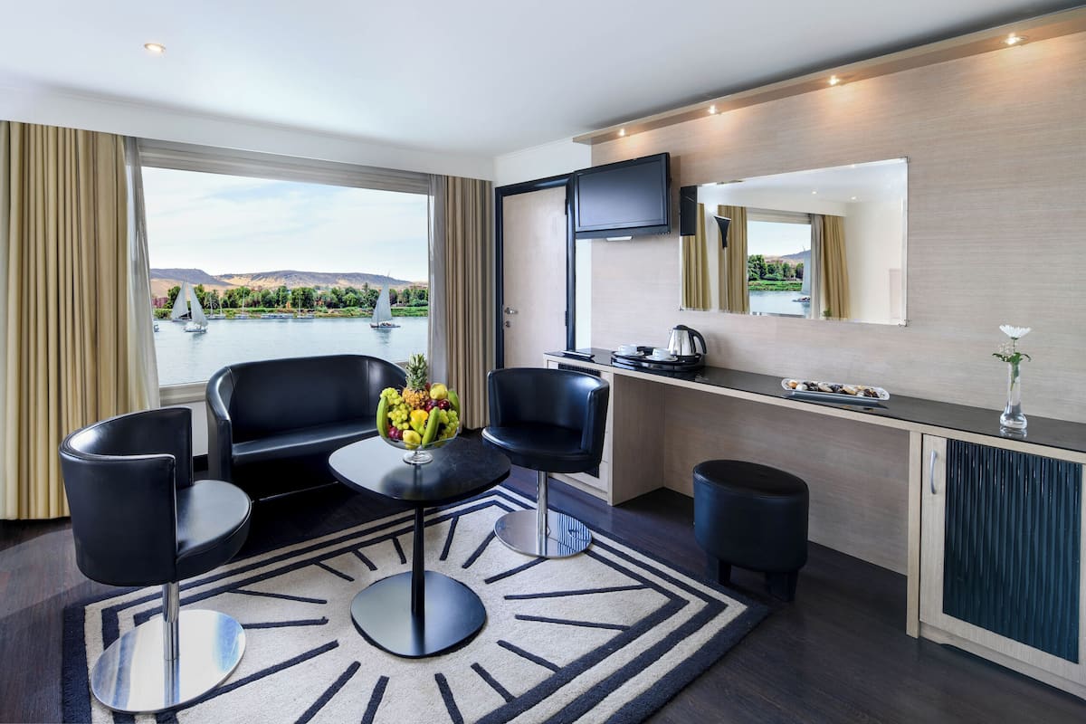 A modern hotel room with a large window, a dining area, a flat-screen TV, and a view of a lake with sailboats.
