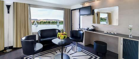 Habitación de hotel moderna con un ventanal grande, zona de comedor, televisor de pantalla plana y vistas a un lago con veleros.