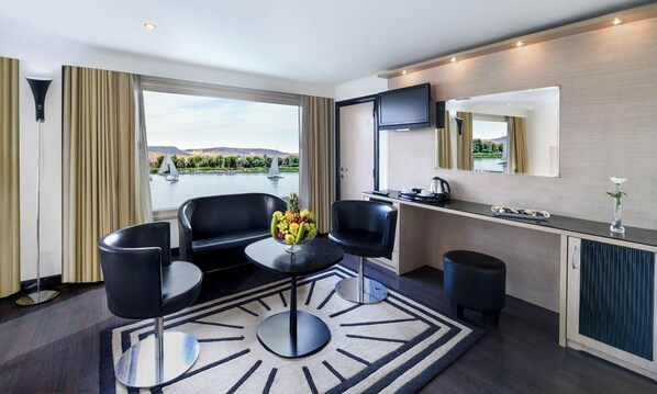 A modern hotel room with a large window, a dining area, a flat-screen TV, and a view of a lake with sailboats. - HS Moondance from Luxor( Sat-Wed),or Aswan (Wed-Sat) (Luxor)