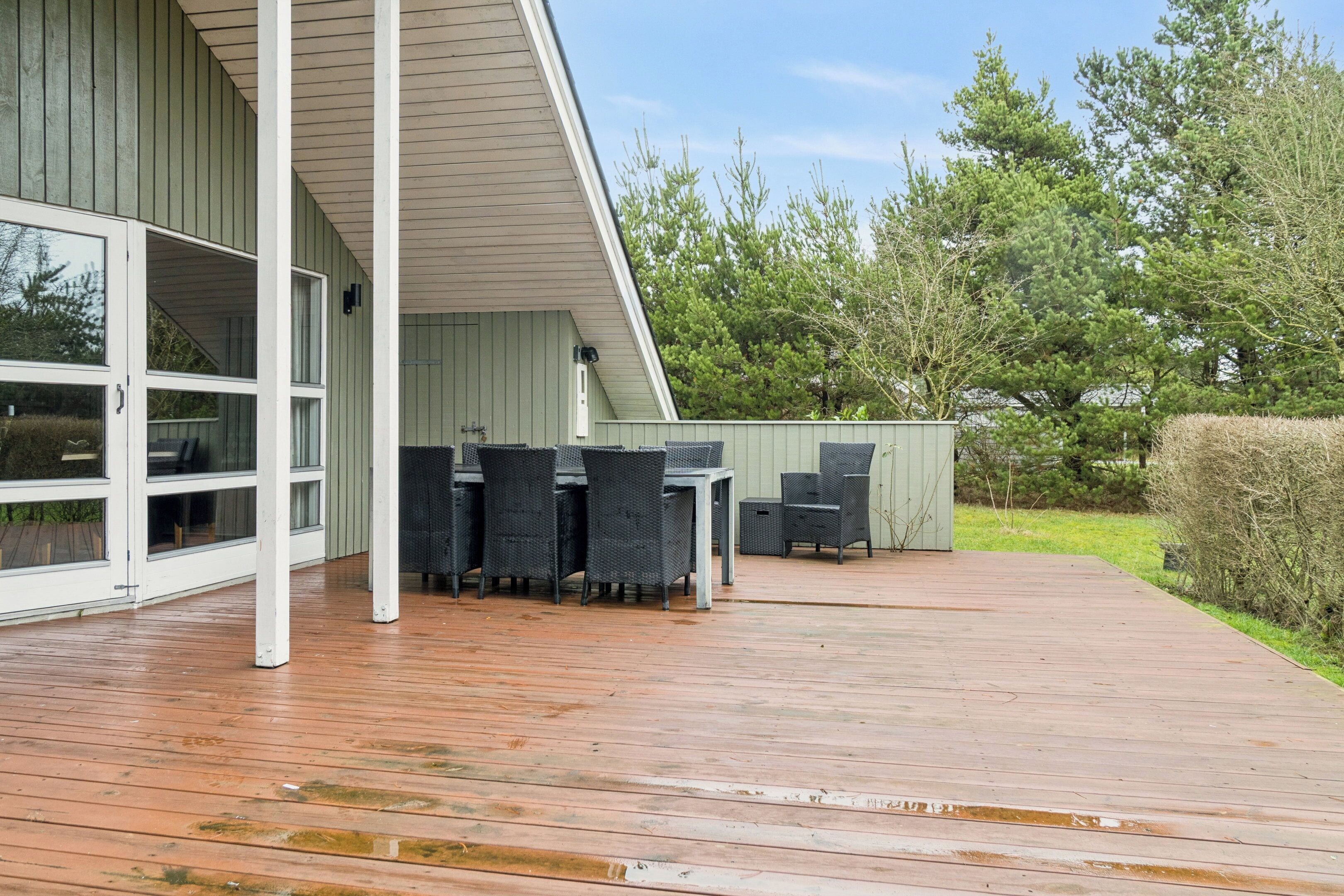 A covered outdoor patio with a dining table and chairs, a white exterior wall, and a wooden deck.
