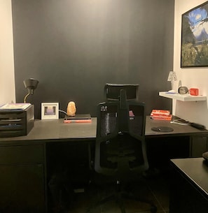 Un bureau de couleur sombre, accompagné d’une chaise de bureau noire, d’une lampe, d’une horloge et de quelques livres.