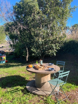 Une table ronde en bois, entourée de chaises en métal, et le tout est agrémenté de verdure et d’arbres.