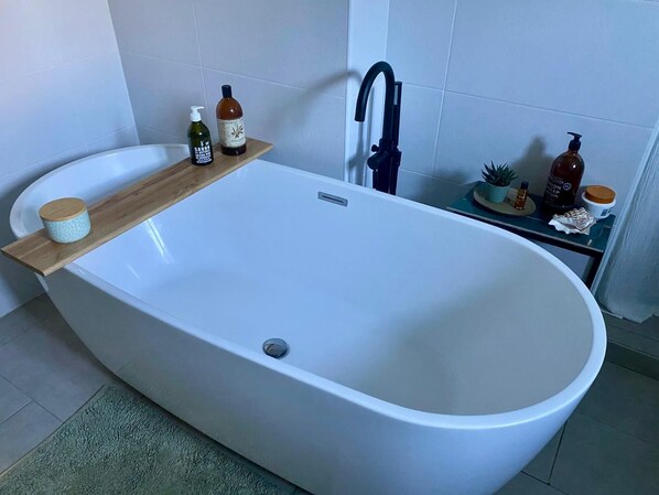 Un baño moderno con una bañera independiente, un estante de madera con artículos de aseo y una mesita con una planta y una botella.
