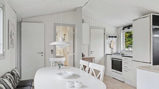 Et lyst interiør med et rundt spisebord, hvide stole, en bænk, et køkken med hvide skabe og et vindue.