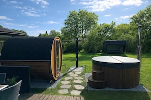 Un bain à remous en bois, une cuve en bois et une petite structure en bois dans un jardin agrémenté d’un chemin de pierres.