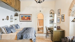 Een gezellige kamer met een bed, een bureau en een stoel. Er hangt een schilderij aan de muur, een kroonluchter en een zicht op een andere kamer door een boog.