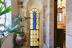 Un arco in pietra conduce a una stanza con un'ampia finestra a vetri colorati, una pianta in vaso e un lampadario.