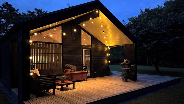 Una cabaña de madera moderna con terraza, asientos exteriores e iluminación colgante.
