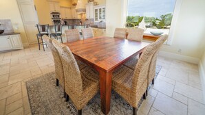 Un comedor con una mesa de madera y sillas de mimbre, una cocina al fondo y un amplio ventanal con vistas al verdor.