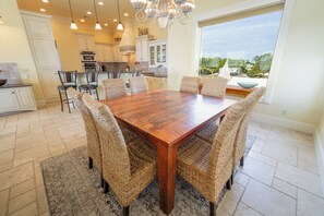 A dining area with a wooden table and wicker chairs, a kitchen in the background, and a large window offering a view of greenery. - August 2026 Big Savings!!! Large home with private pool, hot tub (Eastpoint)