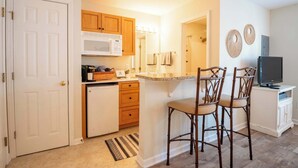 A compact kitchen with wooden cabinets, a microwave, and a small refrigerator. There are bar stools and a television on a cabinet. - The Driftwood: Efficiency Suite in St. James with Pool! (Southport)