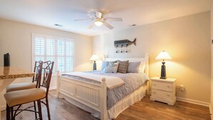 A bedroom with a bed, two bedside tables with lamps, a chair, a window with blinds, and a ceiling fan. - The Driftwood: Efficiency Suite in St. James with Pool! (Southport)