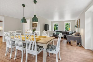 A dining area with a wooden table and white chairs, a gray sofa, and a television in the background. - Cozy country house close to Blokhus and Faarup (Pandrup)