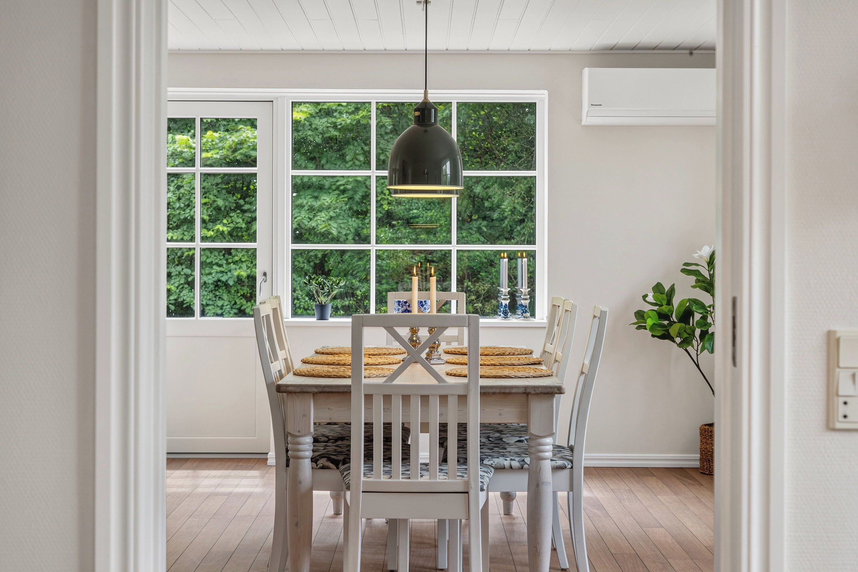 Een eethoek met een tafel en stoelen, een hangende lamp en een raam met uitzicht op groen.
