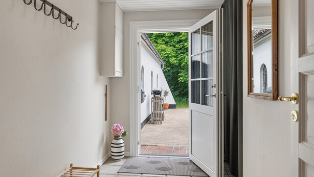 Un couloir avec un porte-manteau en bois, un vase de fleurs, un tapis et une porte ouverte donnant sur une terrasse extérieure.
