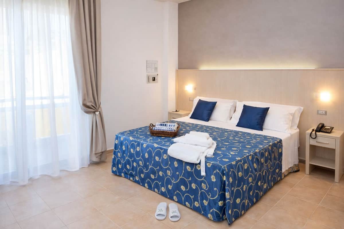 Habitación de hotel con cama, mesita de noche, lámpara, teléfono y ventana con cortinas.
