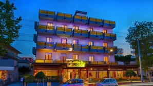 A multi-story hotel with balconies, a neon sign, and a car parked in front. - Hotel Mimosa (Riccione)