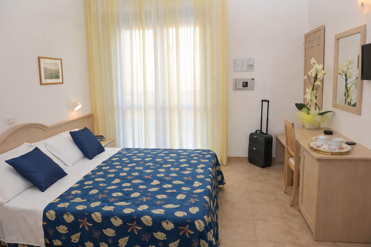 Habitación de hotel con una cama, escritorio, silla, maleta y ventana con cortinas.