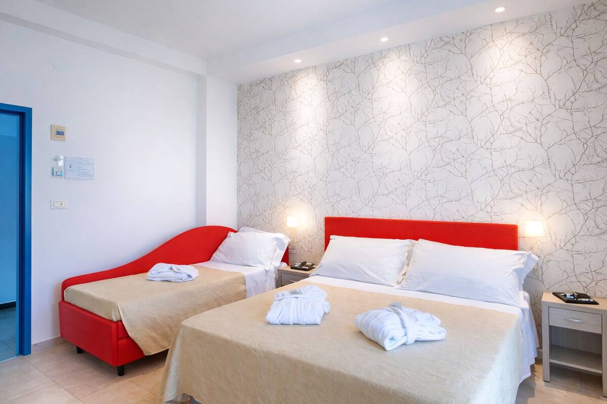 Habitación de hotel con dos camas, cada una con cabecero rojo y ropa de cama blanca. Hay una lámpara de pared sobre cada cama y una mesita de noche pequeña con un teléfono y una lámpara.