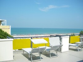 Una terrazza sul tetto con poltrone da lounge bianche e un divisorio giallo, con vista su una spiaggia sotto un cielo azzurro limpido.