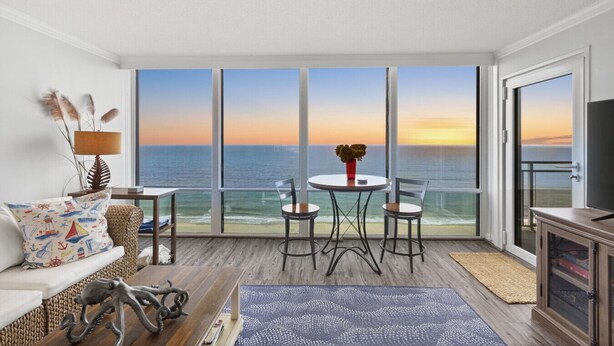 A balcony with a view of the ocean at sunset, furnished with a table and chairs, and a sofa with decorative pillows. - Beachfront Condo/ Pool/ Surfing/ Fishing / EW 1101 (Virginia Beach)
