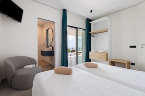 Une chambre d’hôtel avec deux lits, un fauteuil gris, une table d’appoint en bois, un miroir et un téléviseur fixé au mur.