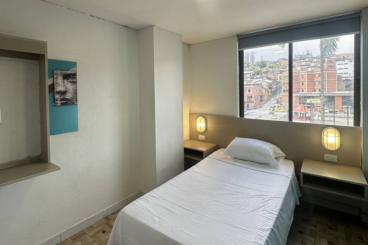 A hotel room with a bed, bedside tables, a window with a city view, and a ceiling light.
