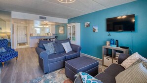 A modern living room with a blue accent wall, a flat-screen TV, and a cozy seating area with a sofa and ottoman. - King`s Getaway Oceanview Pool (Daytona Beach Shores)