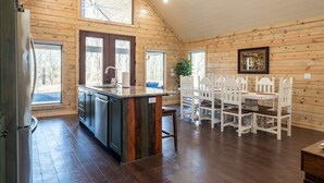 A kitchen with a central island, wooden walls, and a dining area with white chairs. - Riverfront Retreat | Beach, Fishing & Trails (Broken Bow)