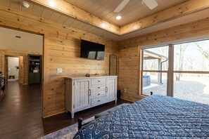 A log cabin bedroom with a bed, a TV mounted on the wall, a wooden cabinet, and a large window.
