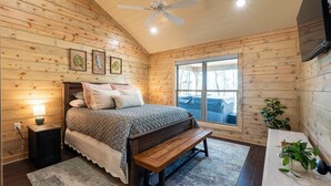 A wooden-paneled bedroom with a bed, bedside lamp, bench, and a window with a view. - Riverfront Retreat | Beach, Fishing & Trails (Broken Bow)