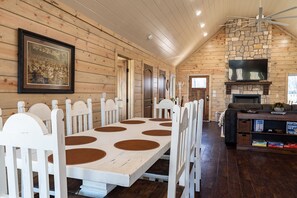 A long dining table with white chairs, a stone fireplace, and a television.