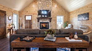 A log cabin interior with a stone fireplace, leather sofa, and a table with books and decorative items. - Riverfront Retreat | Beach, Fishing & Trails (Broken Bow)
