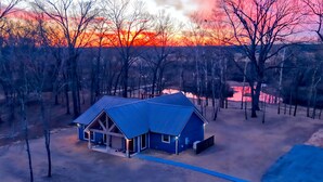 A house with a blue roof, surrounded by trees, and a sunset in the background. - Riverfront Retreat | Beach, Fishing & Trails (Broken Bow)
