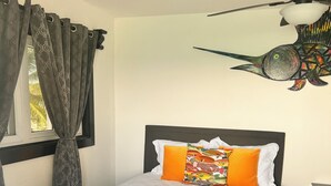 Una cama con una colcha blanca texturizada, un cojín colorido y un ventilador de techo con un adorno de pez.